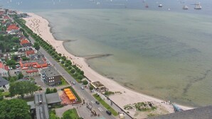 Uma vila costeira com uma praia de areia, uma estrada com carros estacionados e vista para o mar com barcos.