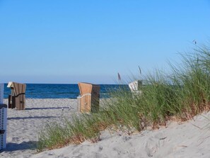 A beach with two wicker chairs and a white chair, surrounded by sand and grass.