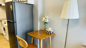 Ein kleines Zimmer mit Holztisch, Stuhl, Stehlampe, Kühlschrank und Waschmaschine.