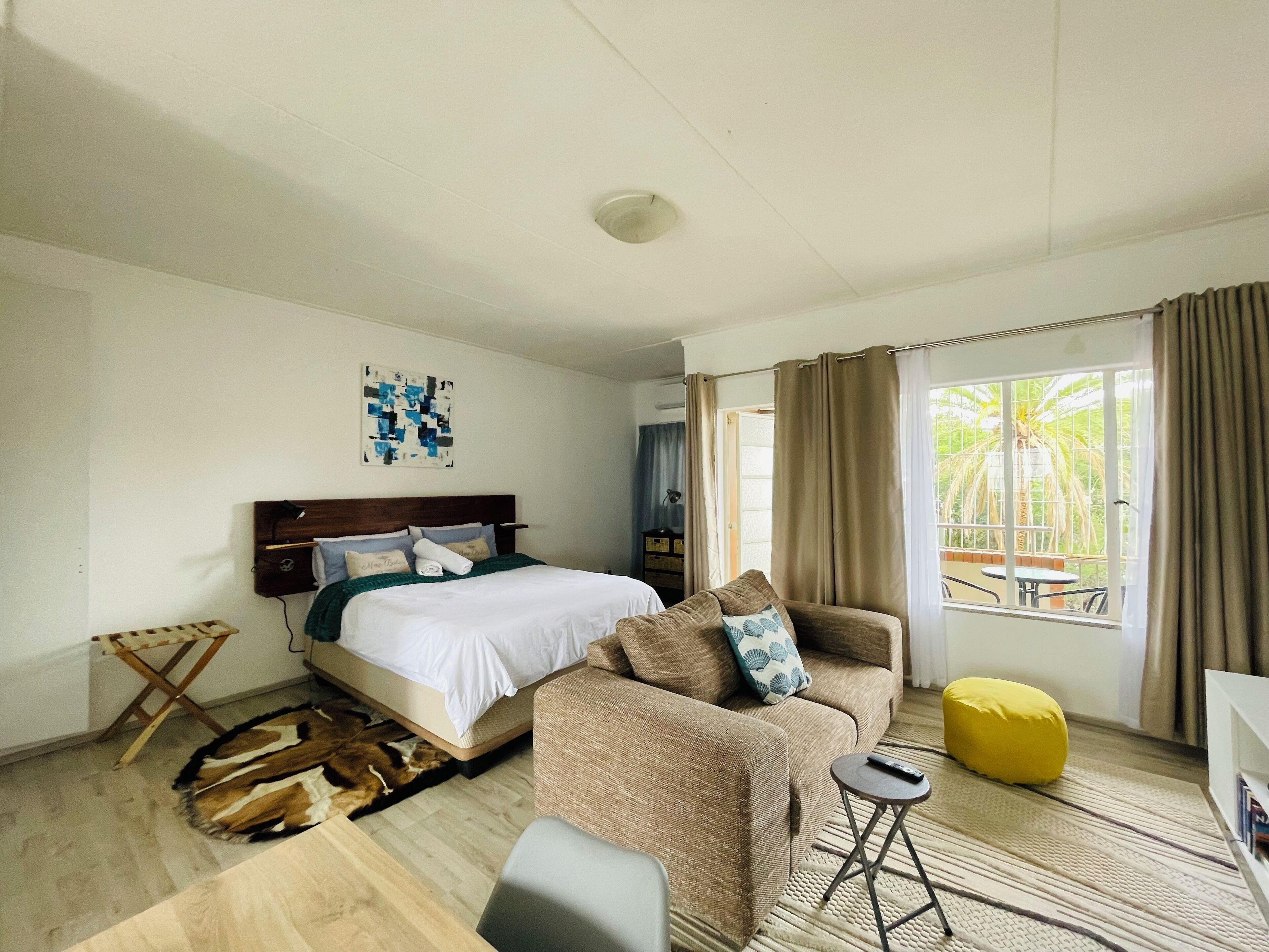 A hotel room with a bed, a sofa, a yellow ottoman, a small side table, and a view of the outdoors.