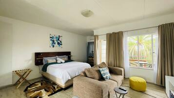 A hotel room with a bed, a sofa, a yellow ottoman, a small side table, and a view of the outdoors.