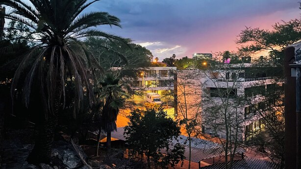 Un edificio de varios pisos con terraza en la azotea, rodeado de árboles y un cielo al atardecer.