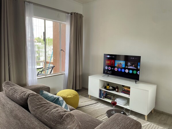 A living room with a flat-screen TV, a white entertainment unit, and a large window with curtains.