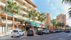 A street with parked cars, palm trees, and multi-story buildings with balconies.