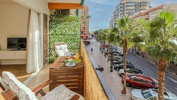 A balcony with a wooden table, a TV, and a view of a street with parked cars and palm trees.