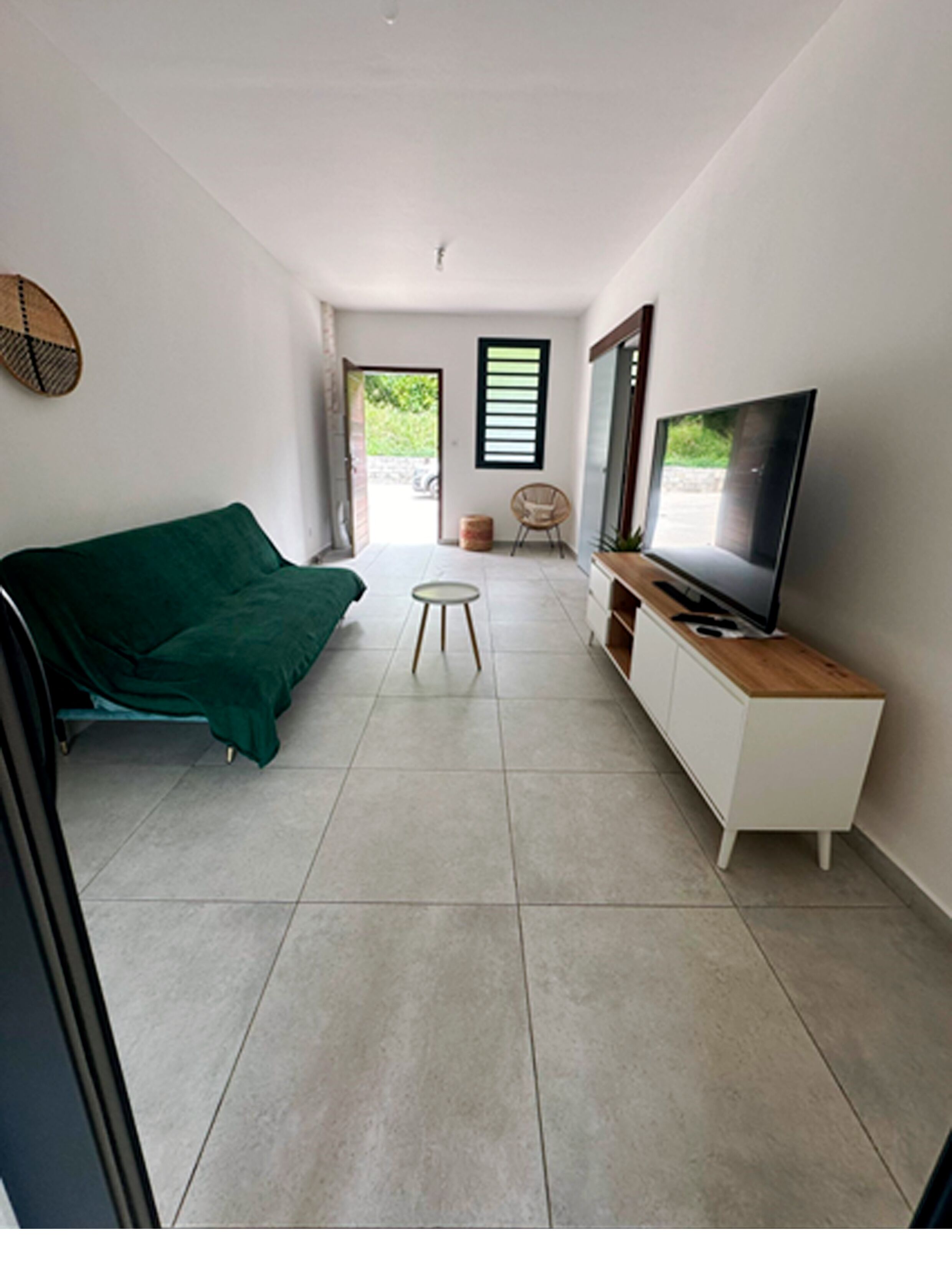 A modern, minimalist living room with a green sofa, a small round table, a TV on a wooden cabinet, and a view of the outdoors through a window.