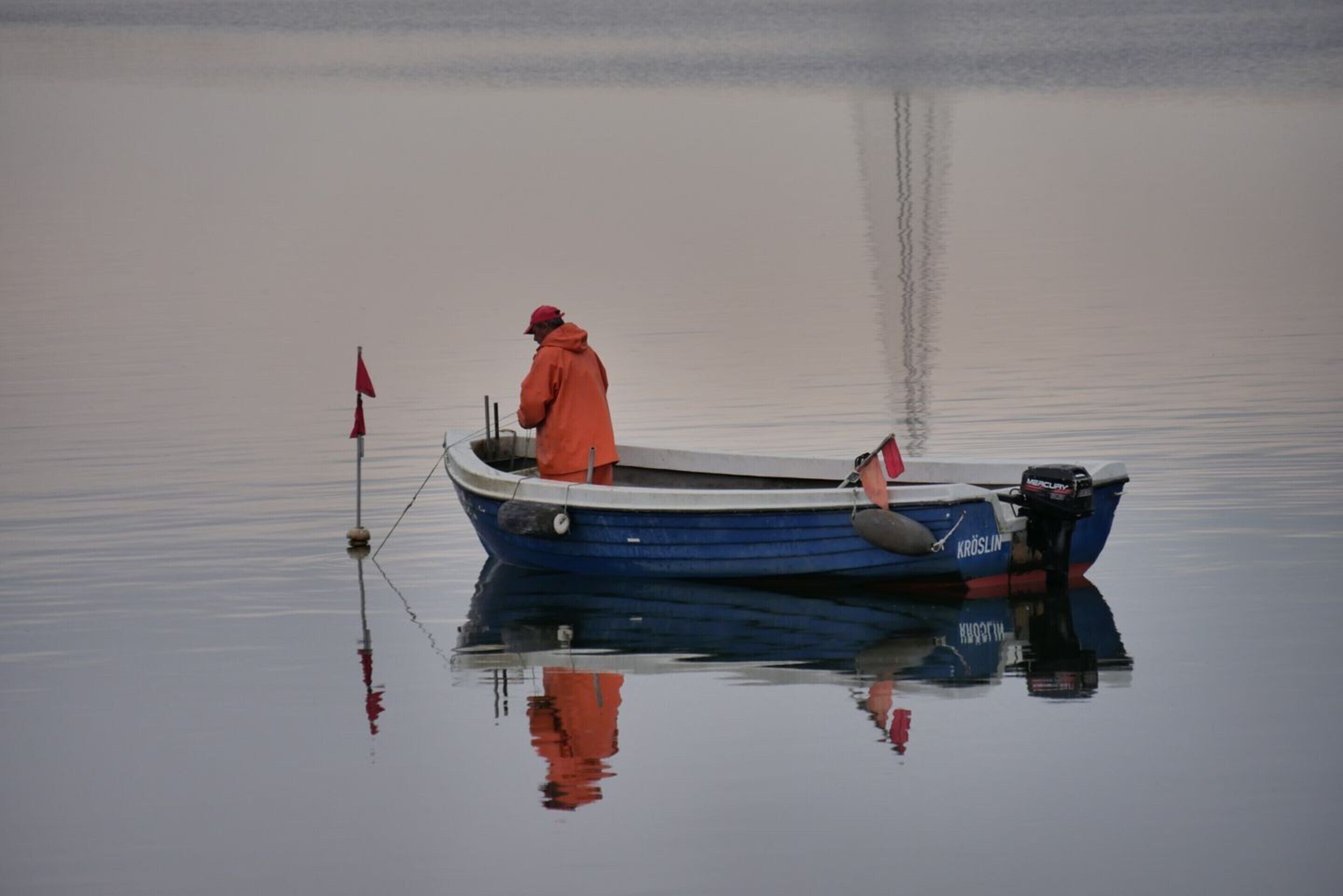 Una persona con una giacca arancione su una piccola barca blu con motore, ancorata vicino a una boa rossa in acque calme.