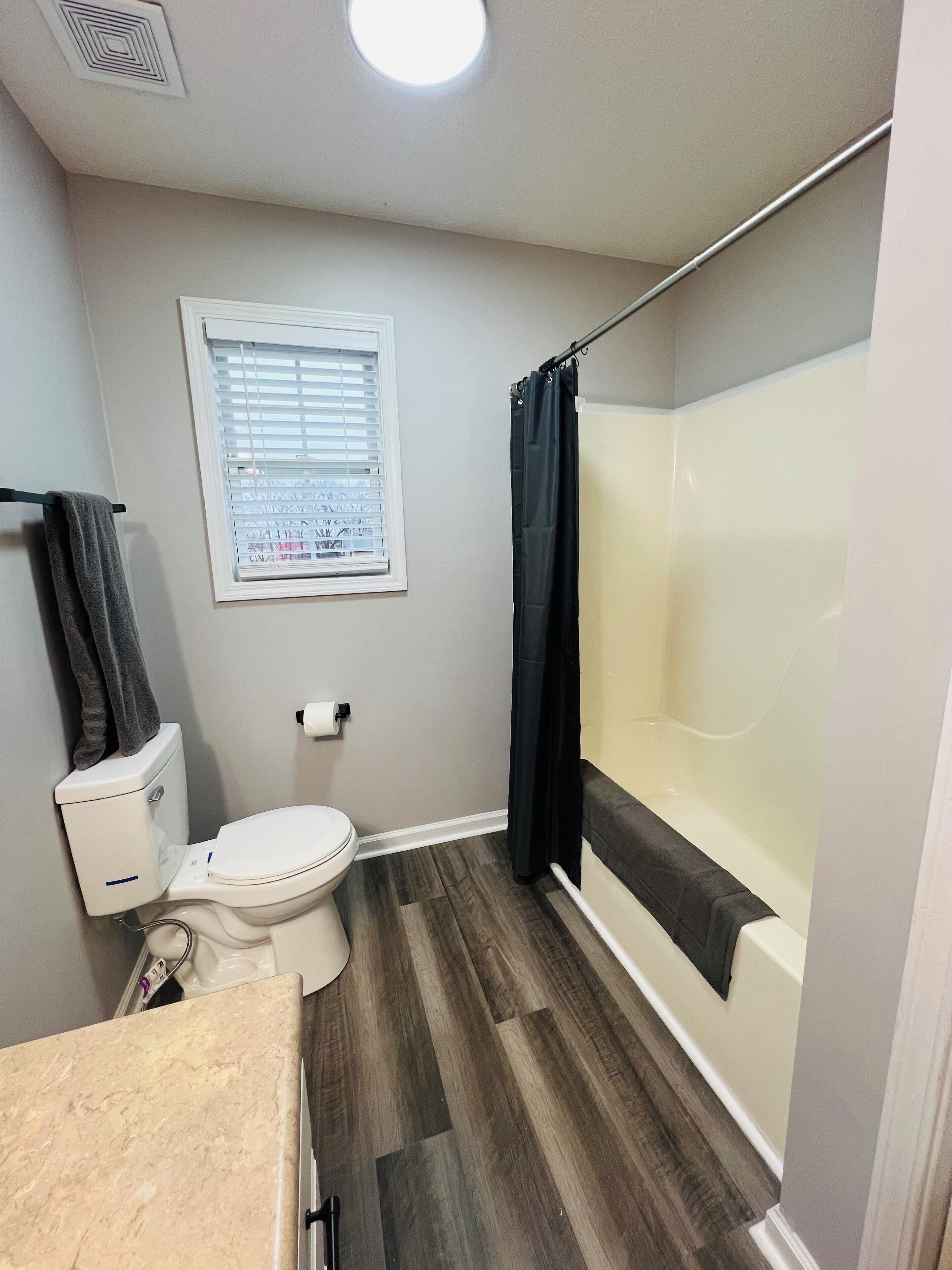 A bathroom with a toilet, a shower with a black curtain, a window with blinds, and a marble countertop.