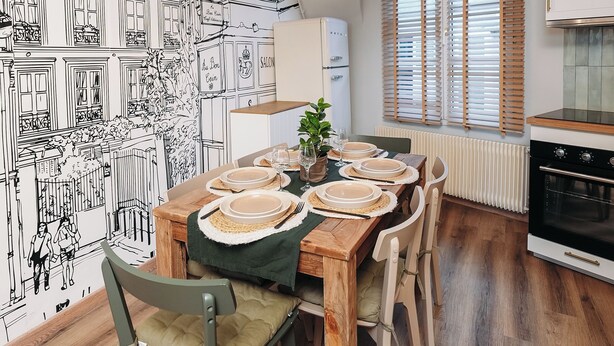 Un coin repas avec une table en bois dressée pour quatre personnes, entourée de chaises dotées de coussins verts. Une grande fresque murale représente un paysage urbain.