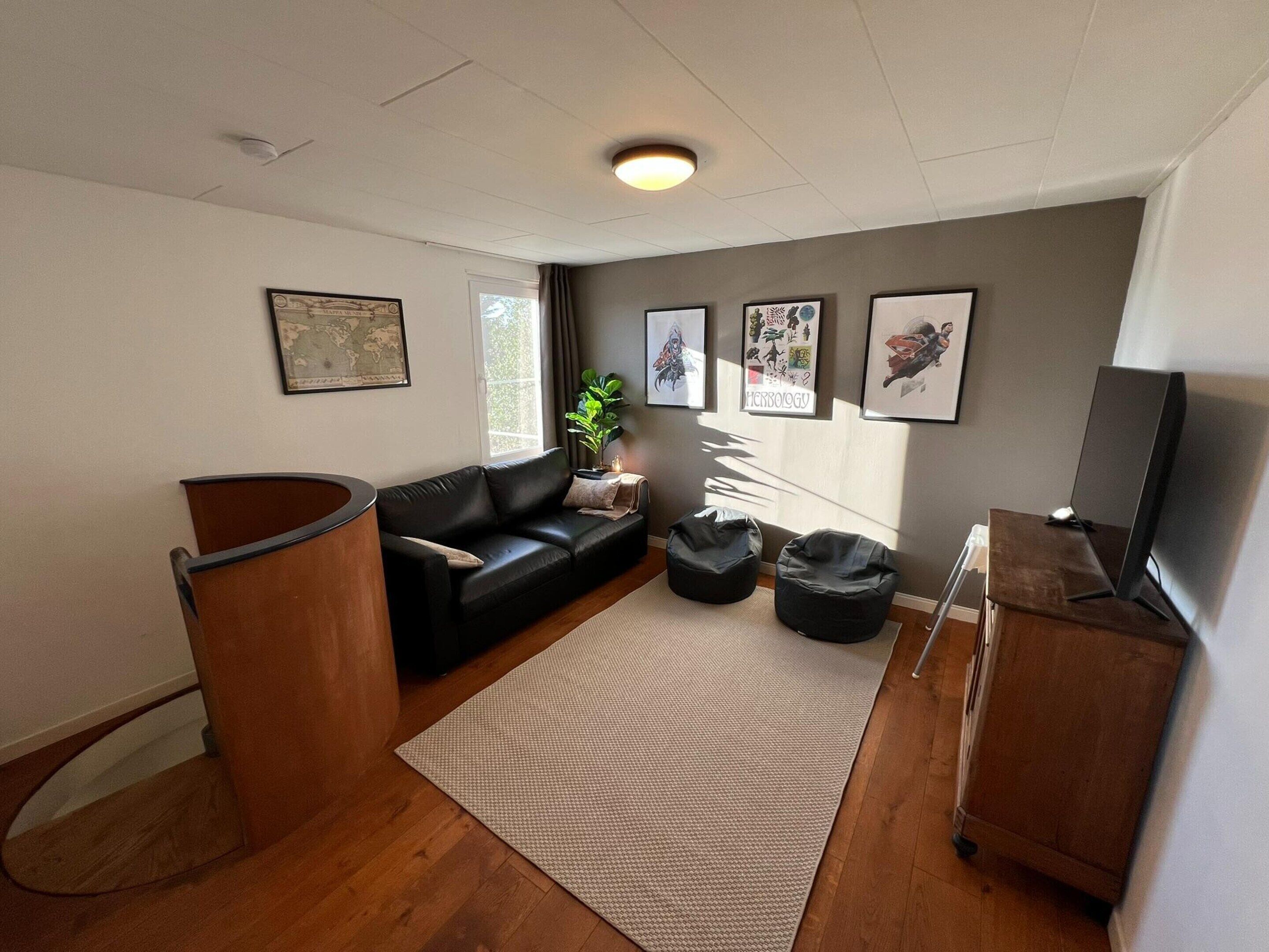 Ein modernes Wohnzimmer mit einem schwarzen Ledersofa, zwei Sitzsäcken in Schwarz, einem Holzschrank und einem Fernseher auf einem Standfuß.