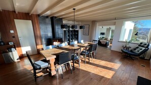 A spacious living area with a wooden floor, a dining table with chairs, a fireplace, and a large window providing a view of the outdoors. - Panoramic Villa With Private Sauna in Erezee (Erezee)