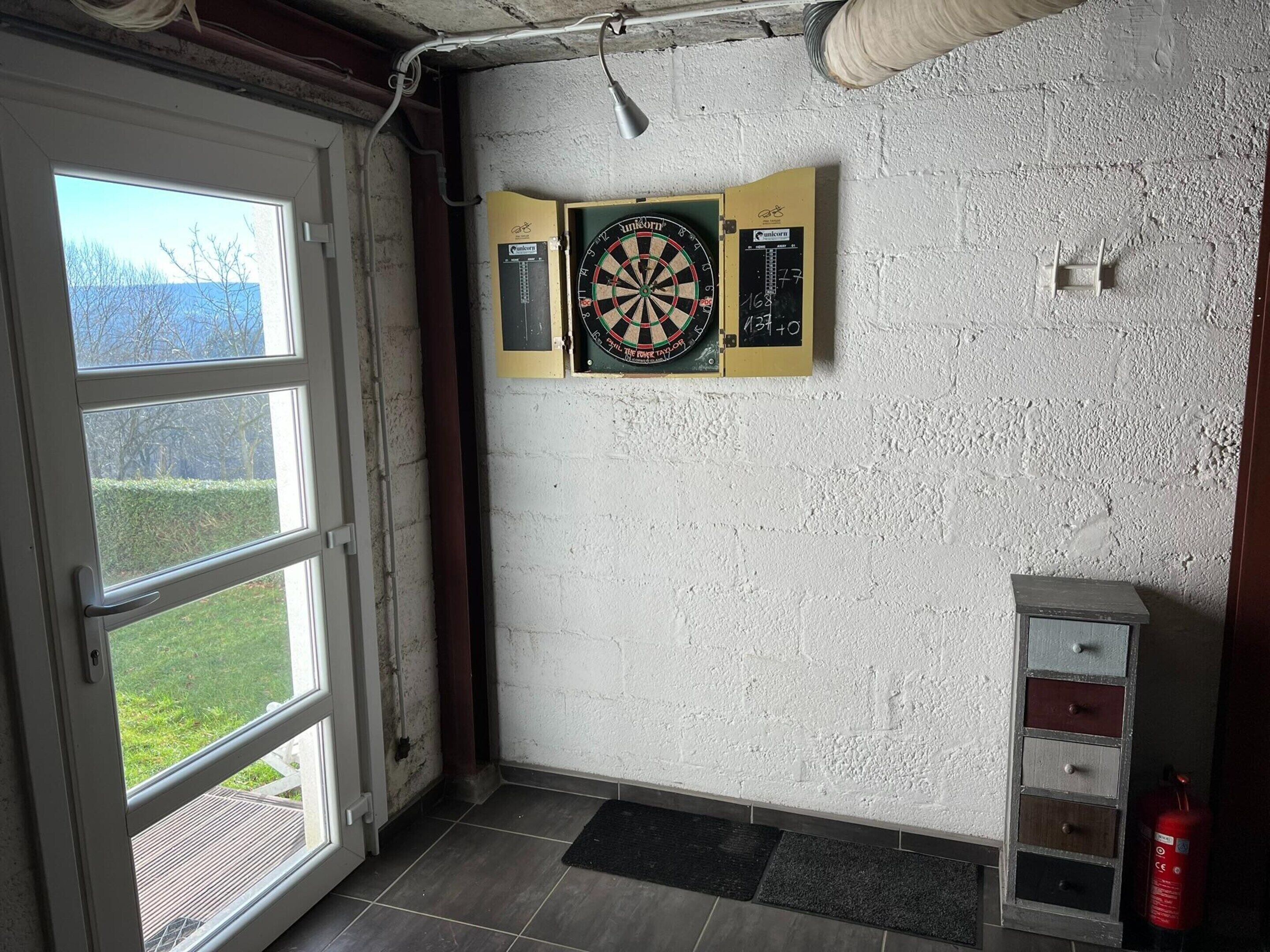 A room with a dartboard, a fire extinguisher, and a window with a view of trees.