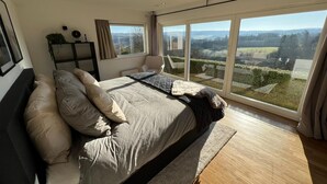 A spacious bedroom with a large bed, a chair, and a view of the countryside through floor-to-ceiling windows. - Panoramic Villa With Private Sauna in Erezee (Erezee)