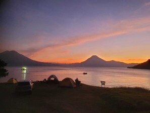 A camping area with tents, a car, and a boat on a lake at sunset with mountains in the background. - Bungalow Bahía Visión Azul (Sololá)
