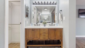 Une salle de bain avec un meuble-lavabo, deux vasques, un miroir avec éclairage intégré, un porte-serviettes et un téléviseur fixé au mur.