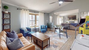 Un vaste espace de vie comprenant un canapé, des fauteuils, une table basse et un baby-foot. On y trouve également un ventilateur de plafond, une cuisine à l’arrière-plan et une grande fenêtre avec des rideaux.