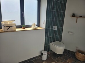Une salle de bain comprenant des toilettes, un bidet, une poubelle, une fenêtre et une étagère avec une plante et un panier.