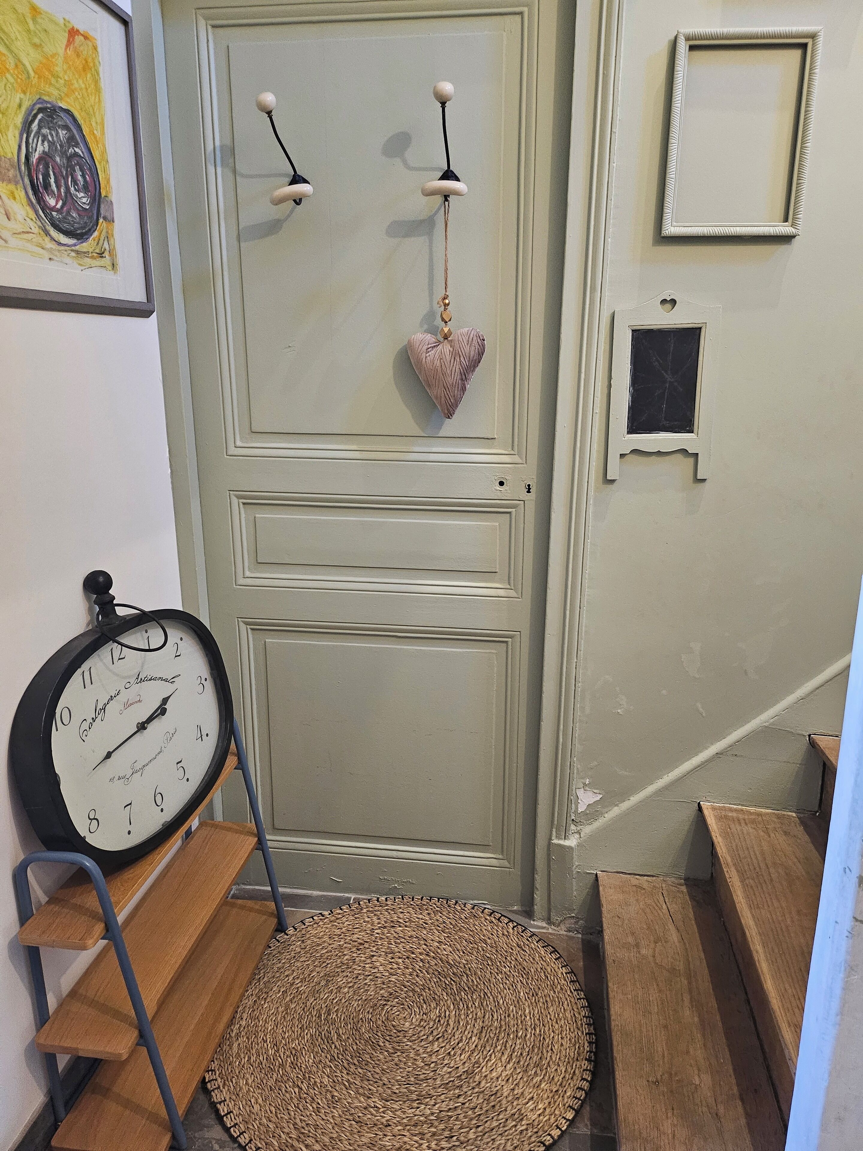 Una escalera con un tapete tejido al pie, un reloj de pared, una decoración con forma de corazón y un ventanal pequeño.