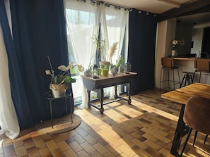 A room with a wooden table, chairs, a potted plant, and a view through a window with curtains. - Nice family cocoon  (Neuilly-Saint-Front)