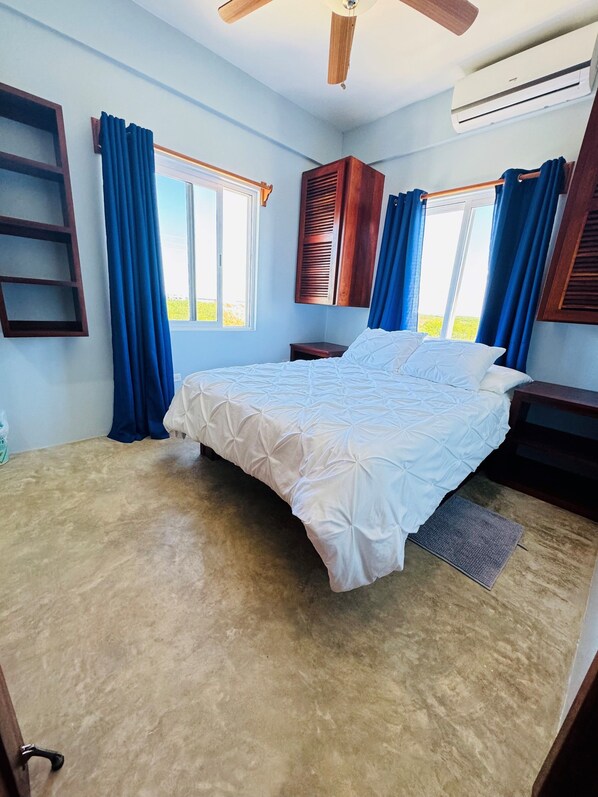 Een slaapkamer met een bed, een plafondventilator, een airconditioning en uitzicht op het landschap door een raam.