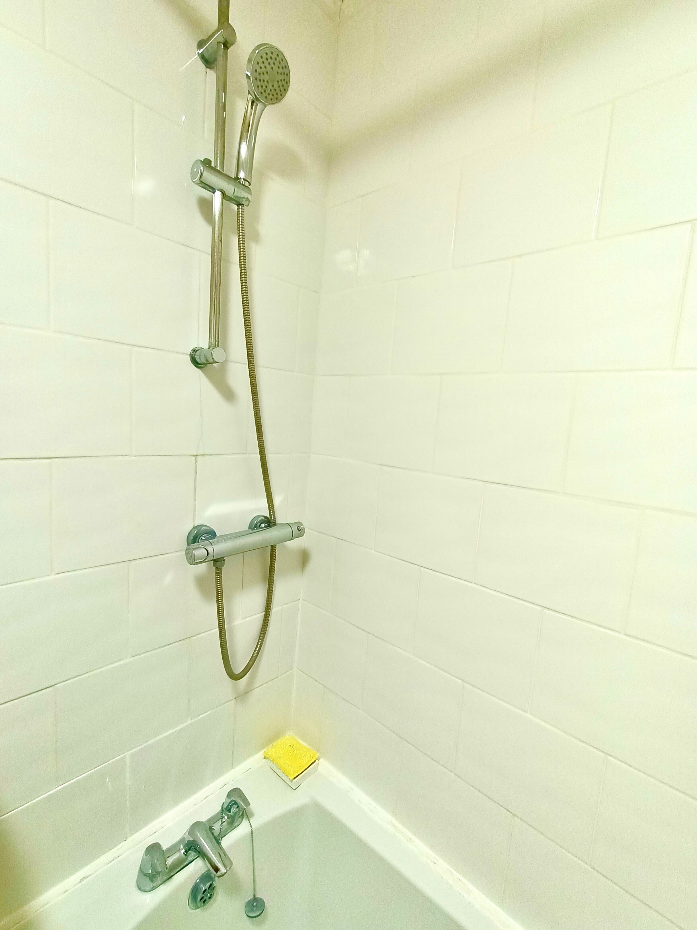 A bathroom with white tiled walls, a shower with a handheld showerhead and a handheld shower attachment, a bathtub with a yellow bath mat, and a faucet.