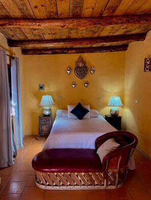 Ein Hotelzimmer mit Holzboden, einem Bett mit weißer Bettwäsche und einem schwarzen Kopfkissen, zwei Nachttischen mit Lampen, einer hölzernen Bank und einem Sessel.