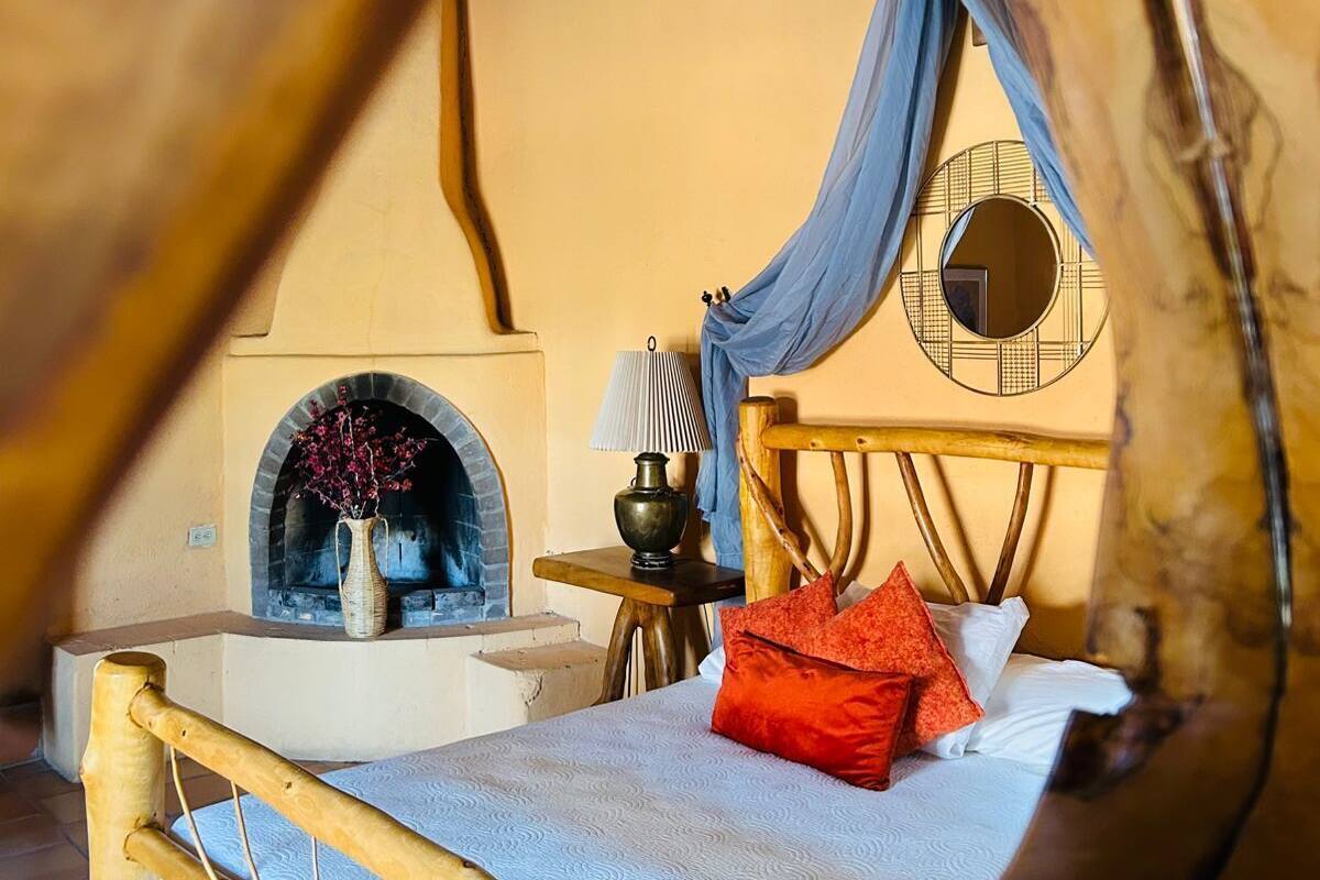Ein gemütliches Zimmer mit Bett, einem kleinen Tisch mit Lampe, einem Kamin und einem Fenster mit Vorhängen.