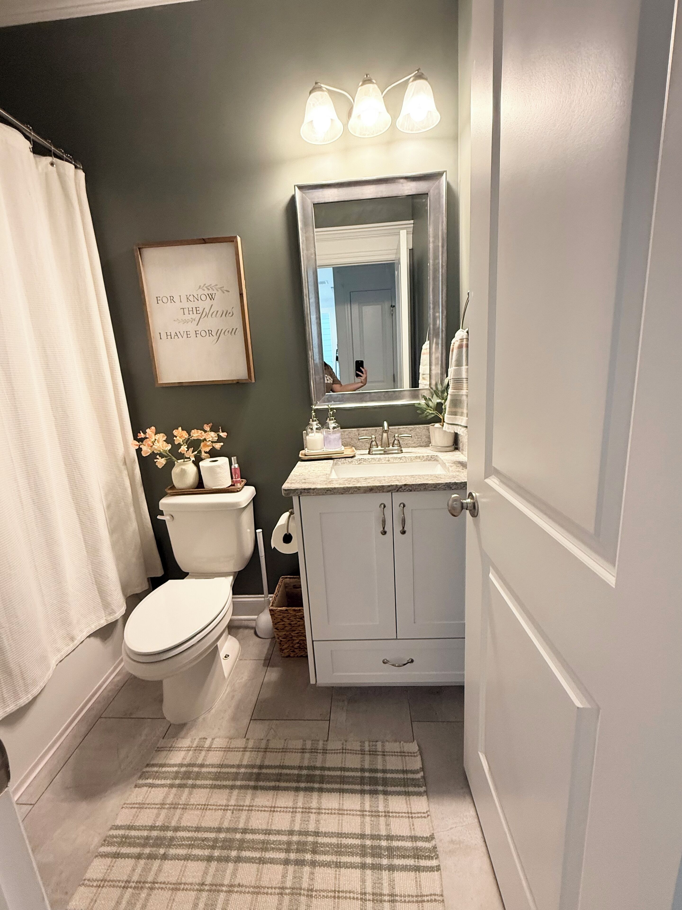 Une salle de bain comprenant des toilettes, un lavabo, un meuble-lavabo et un miroir. Un cadre avec une citation est accroché au mur, et il y a un rideau de douche.