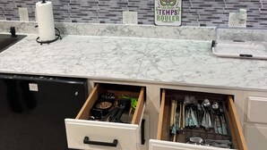 Una cocina con armarios blancos, encimera de mármol y salpicadero de azulejos. Hay dos cajones debajo de la encimera, un dispensador de toallas de papel y un cartel en la pared.