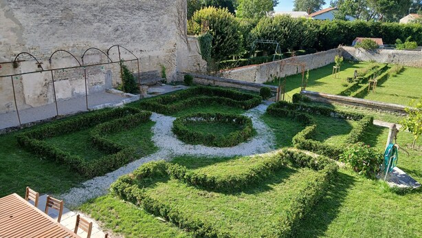 Un cortile con un muro in pietra, un tavolo di legno e un giardino con siepi a geometria.