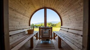 A wooden sauna with benches and a central heating unit, surrounded by a semi-circular wooden structure with a view of a green landscape. - Luxury Lodge in NH: Ski, Hottub, Sauna, Pool Table, Playscape, Game Room & Hike (Jefferson)
