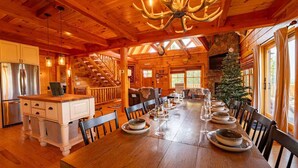 A wooden cabin interior with a dining table set for six, a kitchen with modern appliances, a staircase, and a stone fireplace. - Luxury Lodge in NH: Ski, Hottub, Sauna, Pool Table, Playscape, Game Room & Hike (Jefferson)
