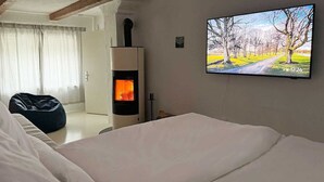A bedroom with a bed, a wall-mounted TV displaying a landscape, and a wood-burning stove. - Cozy Bavarian home with fireplace and fully equipped kitchen. (Moorenweis)