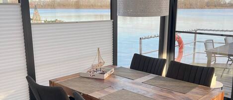 Un comedor con una mesa y sillas de madera, una lámpara colgante, y vistas a un lago y árboles a través de amplios ventanales.