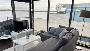 A modern living room with a large gray sofa, a white coffee table, and a flat-screen TV. The room has large windows offering a view of a body of water and a building. - Hausboot am Xantener Nordsee (Xanten)