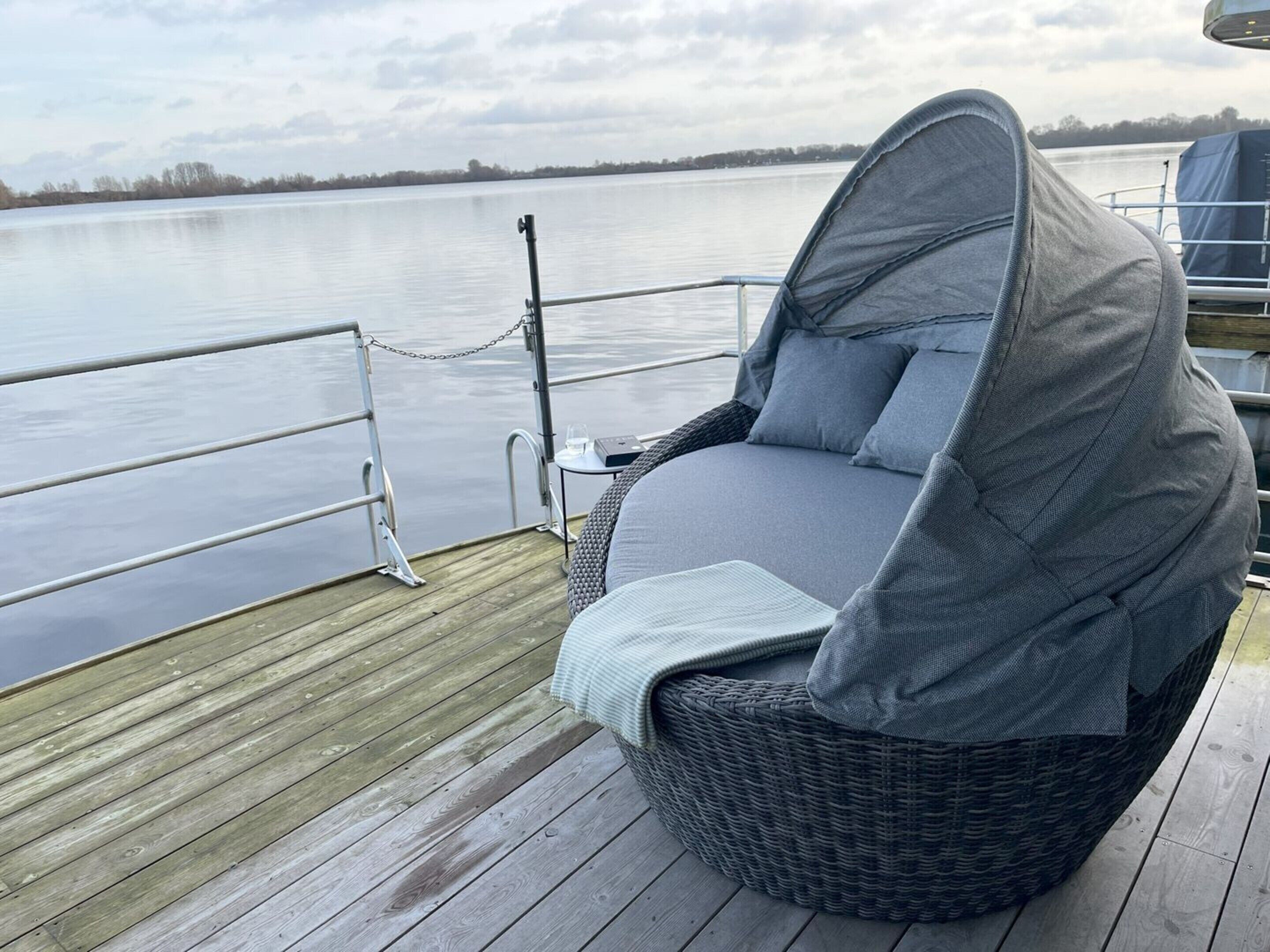 A wicker chair with a grey cushion on a wooden deck by a calm lake.