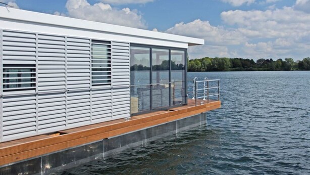 A modern floating house with a wooden deck, large windows, and a view of a lake and trees. - Hausboot am Xantener Nordsee (Xanten)