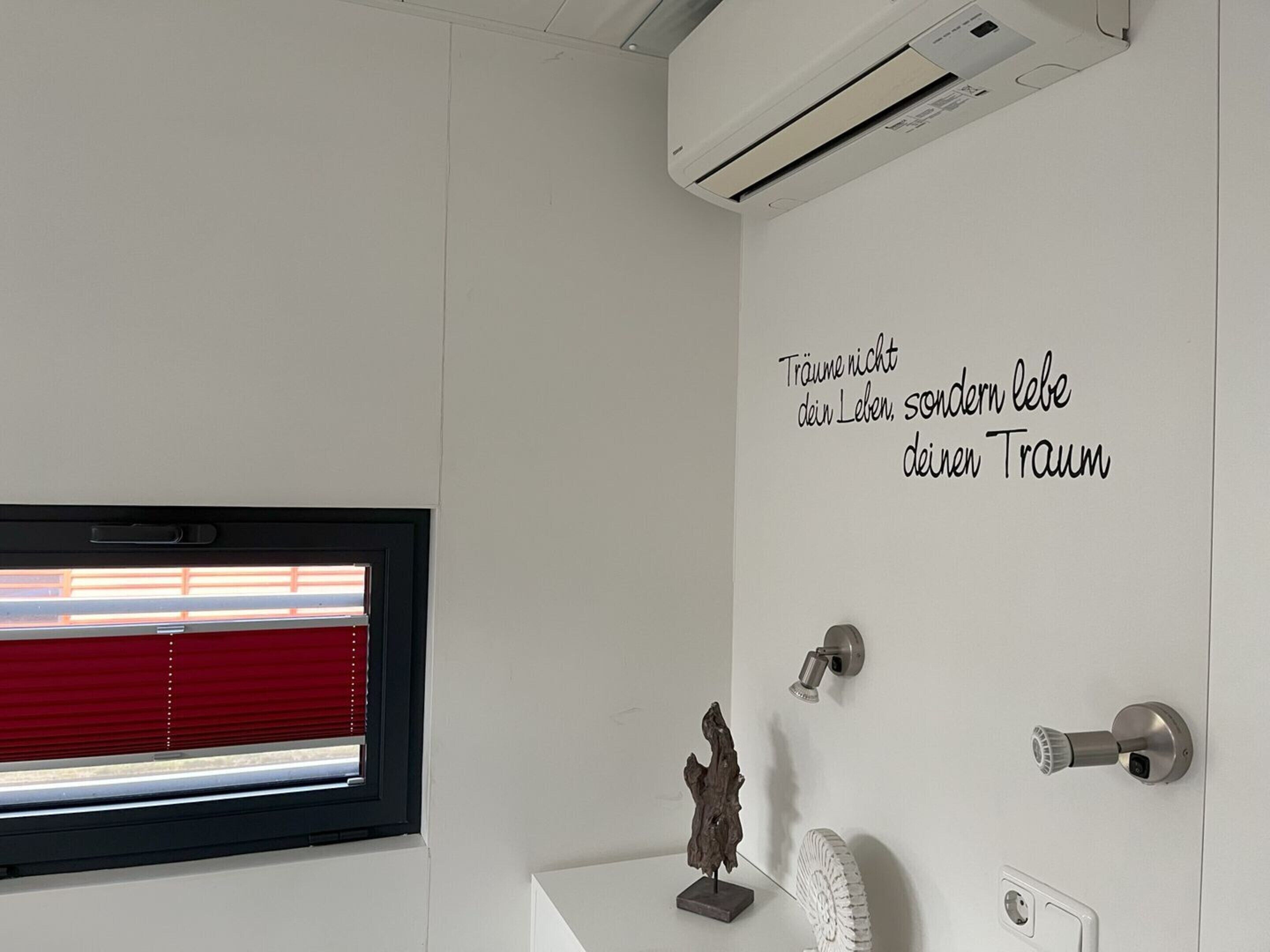 A bathroom with a white wall, a window with red blinds, a wall-mounted air conditioner, a wall-mounted light, a door with a silver handle, and a small sculpture on a pedestal.