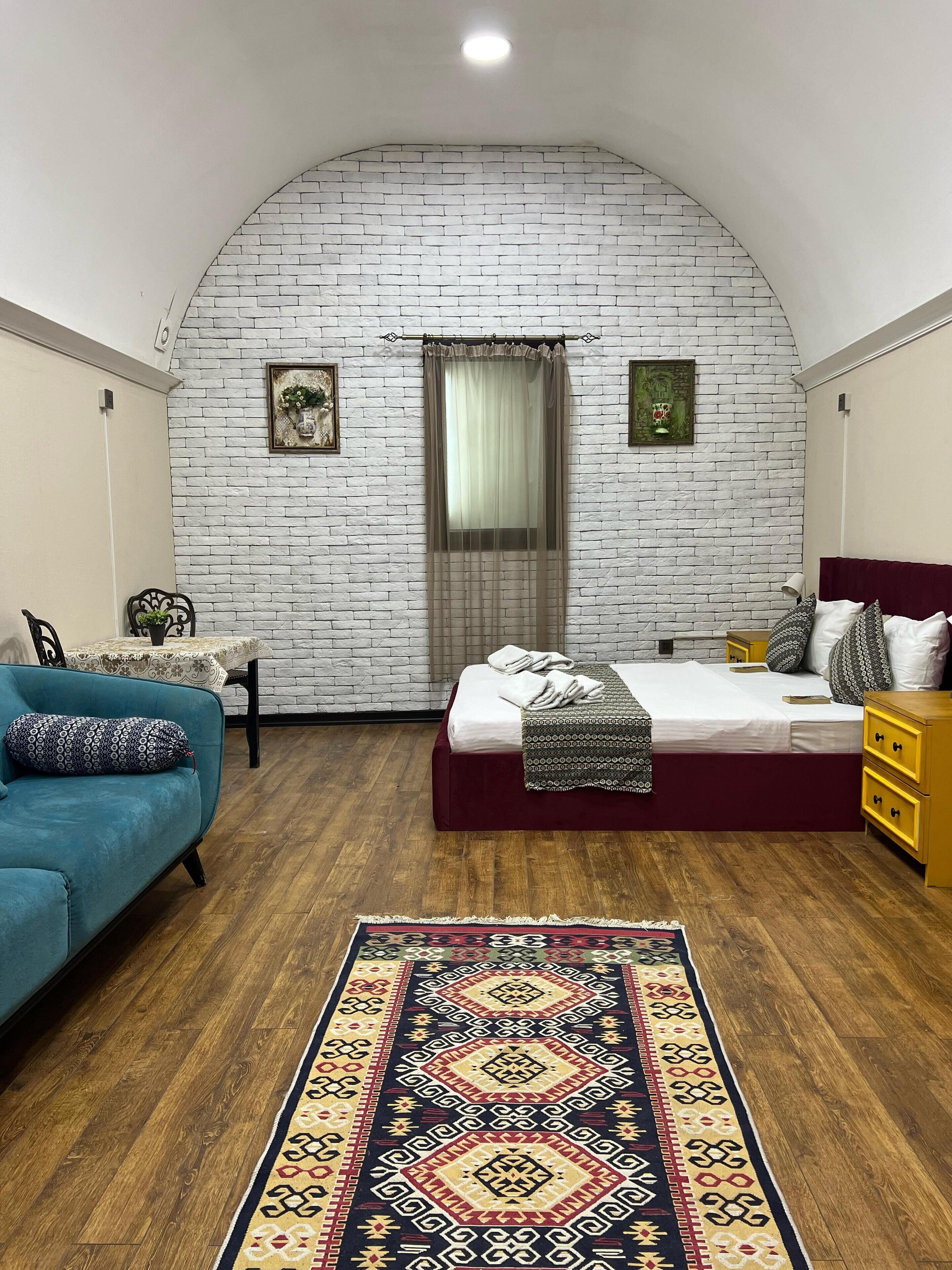 Ein Schlafzimmer mit weißer Ziegelwand, einem Bett mit rotem Kopfteil, einem blauen Sofa, einem gemusterten Teppich und Holzboden.
