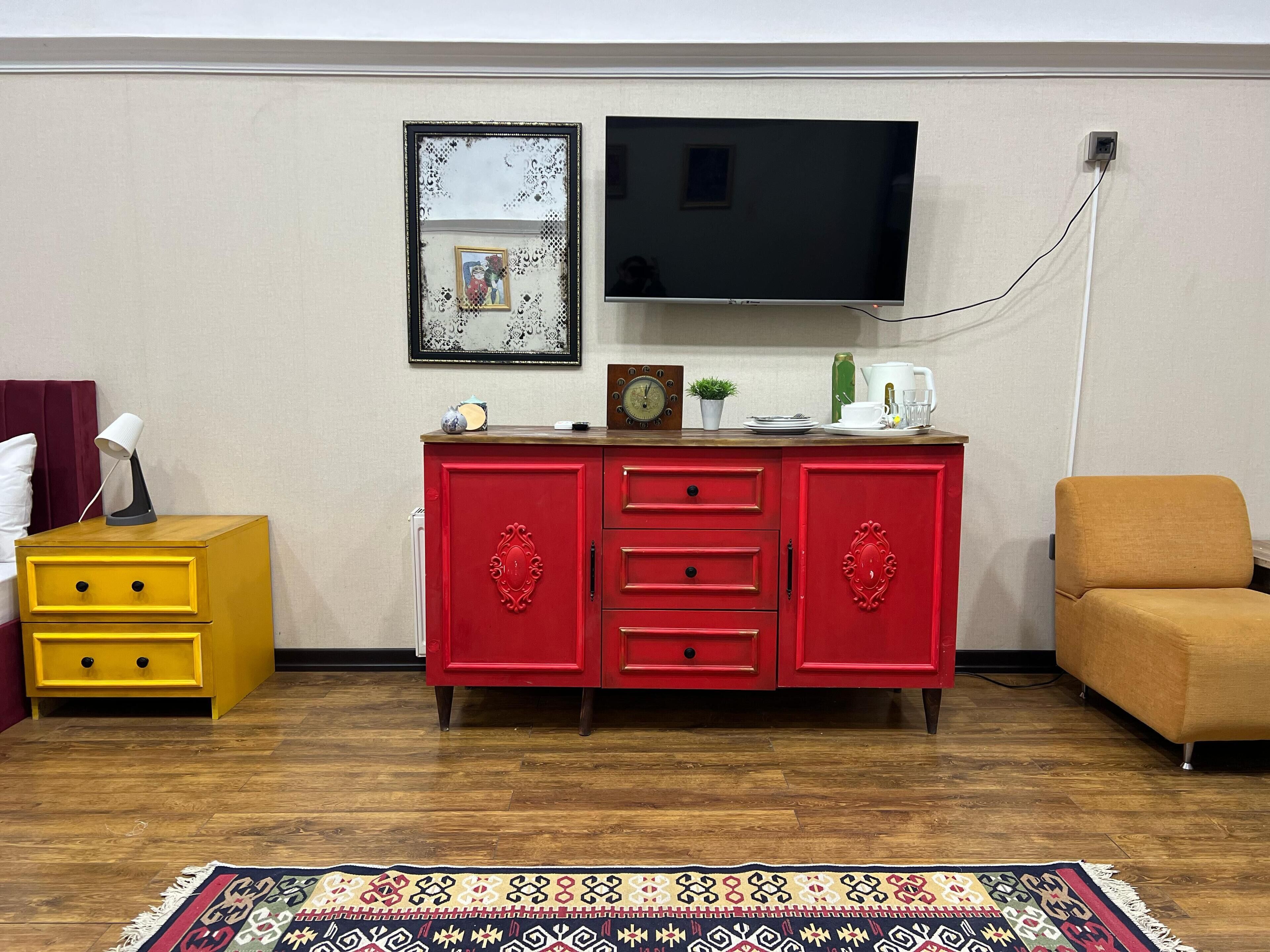 Una sala de estar con piso de madera, un aparador rojo con cajones, una mesita de noche amarilla, un sofá marrón, un televisor de pantalla plana montado en la pared y una alfombra estampada.