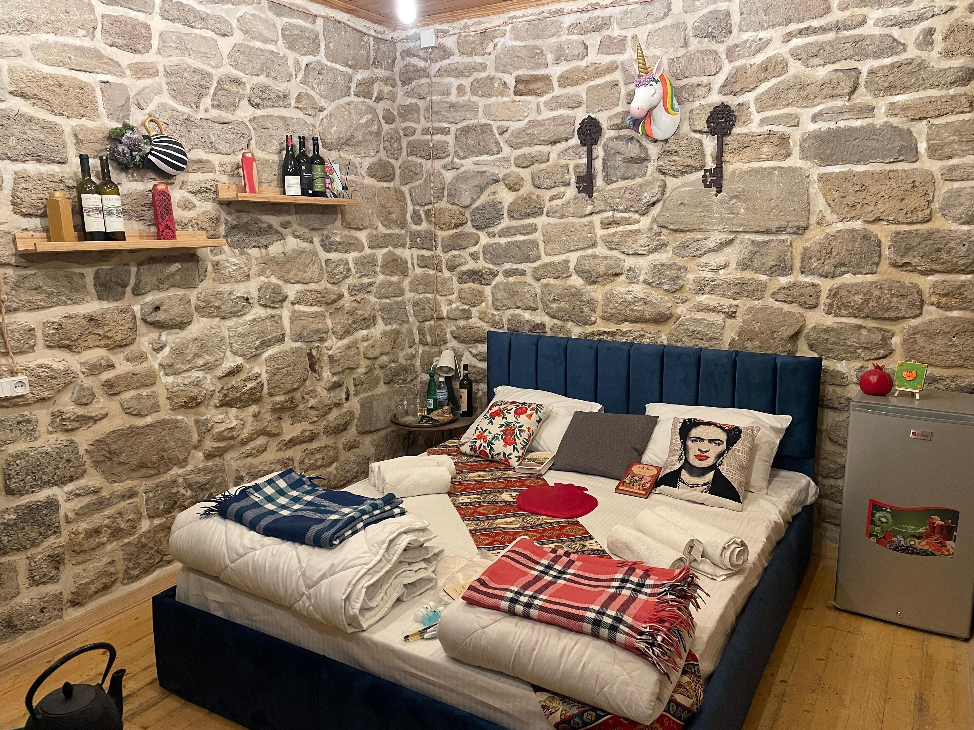 A bedroom with a stone wall, a bed with multiple pillows and blankets, a small table with bottles, and a decorative unicorn wall piece.