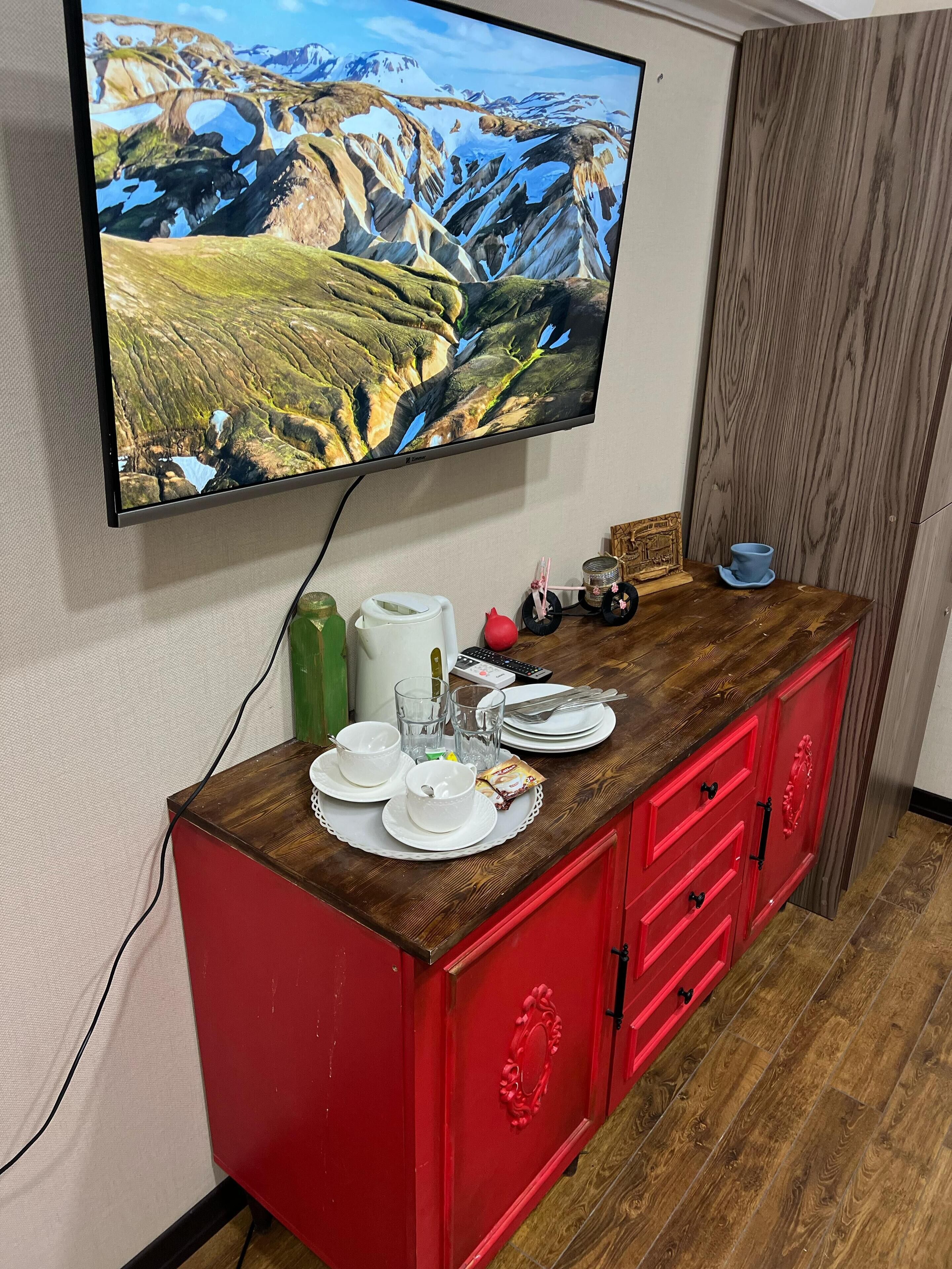 A room with a TV mounted on the wall displaying a mountainous landscape. A wooden table with a red cabinet below holds plates, cups, and a remote control.
