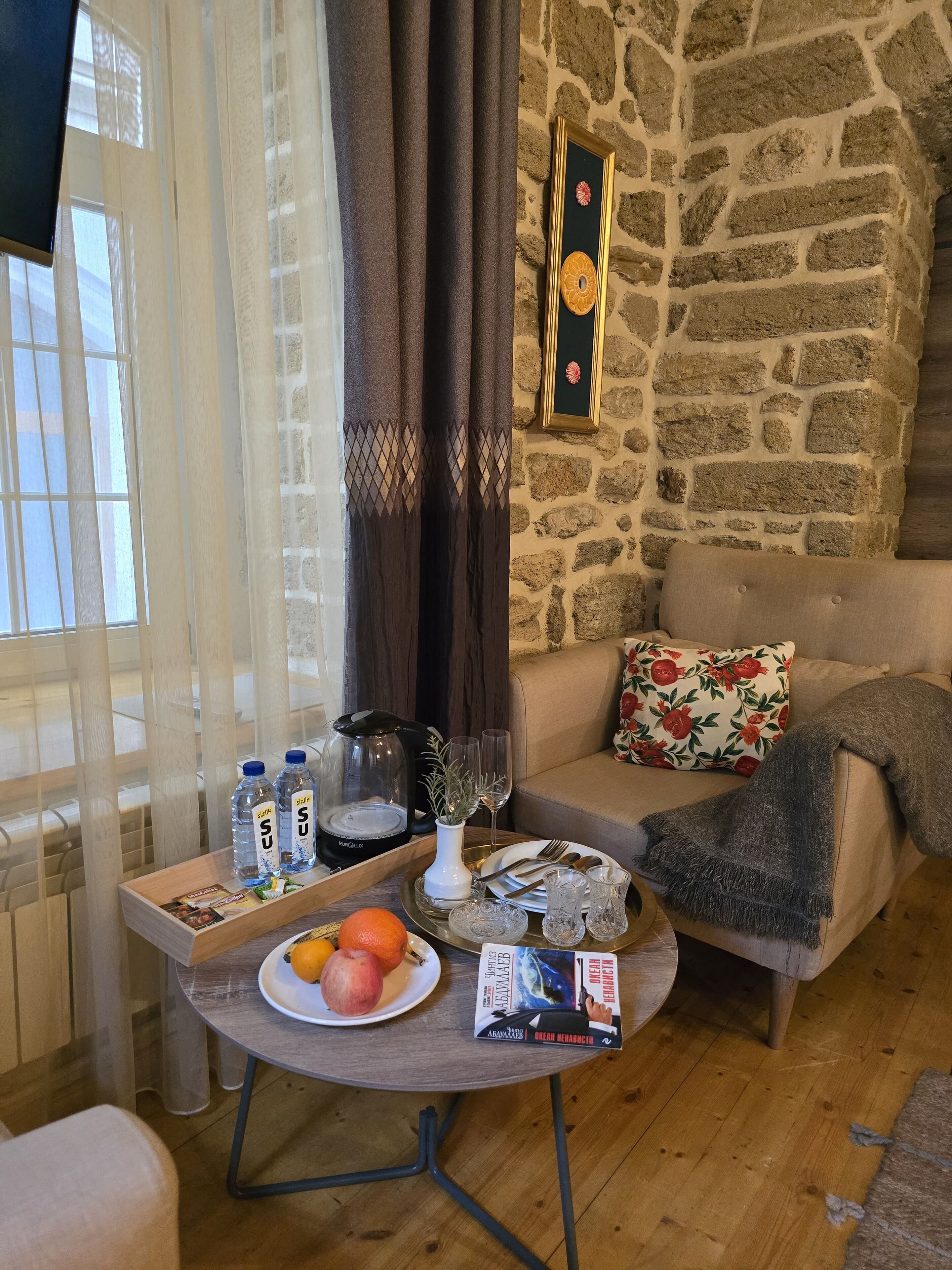 A cozy living area with a stone wall, a sofa with floral cushions, a coffee table with a tray of snacks, a plate with fruits, and a bookshelf with books.