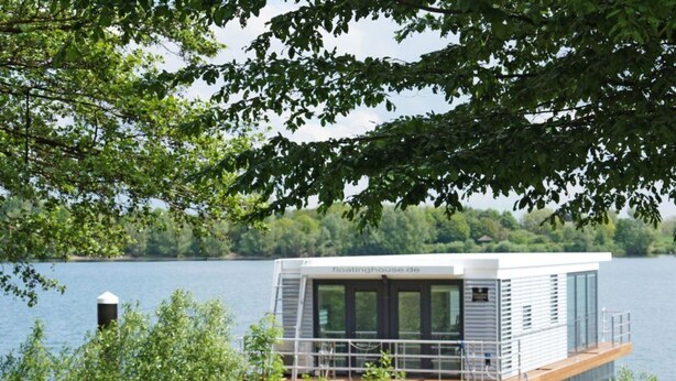 Ein modernes schwimmendes Haus mit Glasfassade, einer Terrasse und Blick auf einen von Grünflächen umgebenen See.