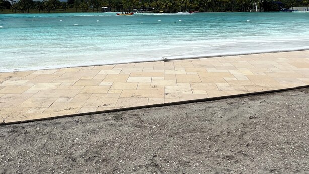 A beach with clear turquoise water, a paved walkway, and palm trees in the background.