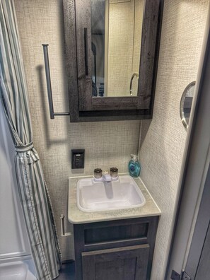 A bathroom with a white sink, dark wooden cabinet, and a mirror above it. There is a striped curtain on the left and a bottle of hand soap on the counter. - Inlet Life Inn - Walk To Everything - (Murrells Inlet)
