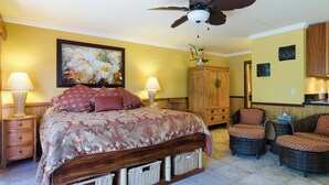 A bedroom with a bed, two bedside tables, a ceiling fan, a wooden cabinet, wicker chairs, and a dining area. - Cozy  studio condo with AC and WiFi in charming Koloa/ Poipu (Koloa)