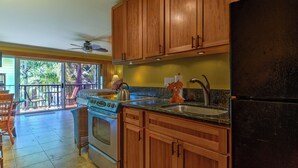 A kitchen with wooden cabinets, a stove, a refrigerator, and a window overlooking a balcony. - Cozy  studio condo with AC and WiFi in charming Koloa/ Poipu (Koloa)