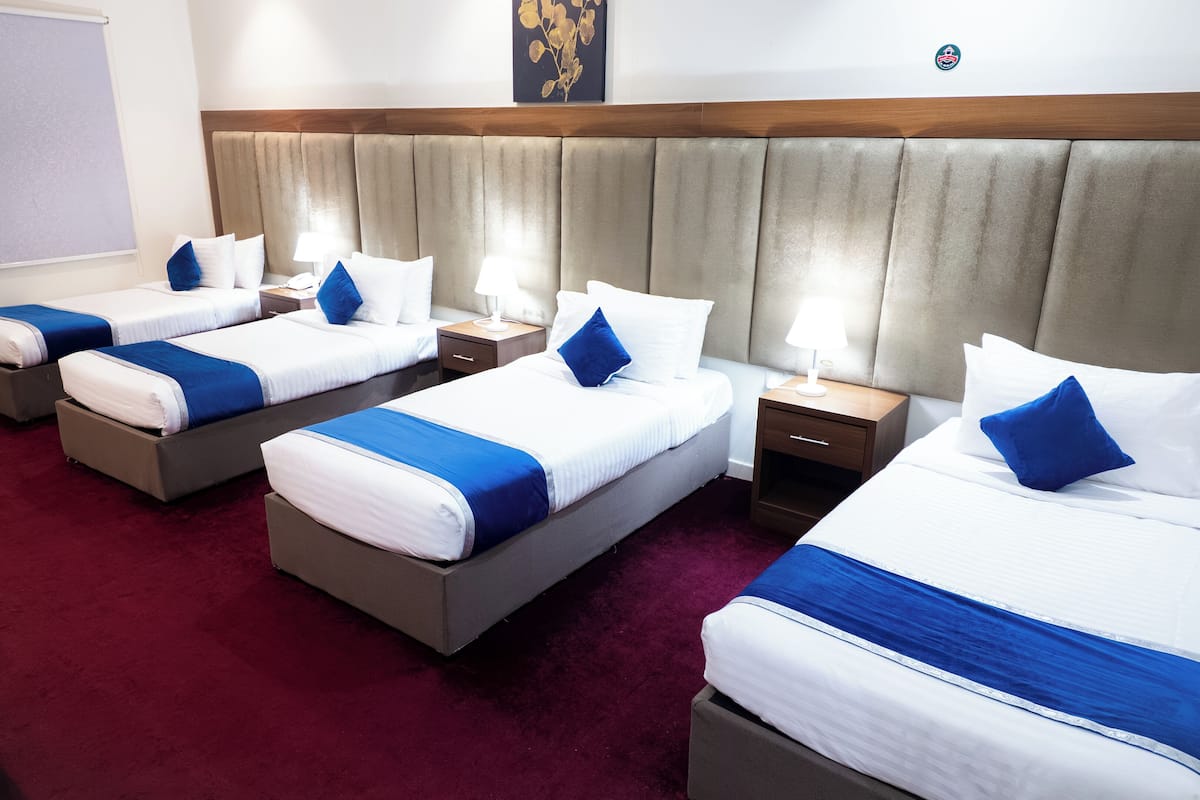 A hotel room with four beds arranged in a row, each with white bedding and blue accents. There are bedside tables with lamps, a wooden headboard, and a red carpet.