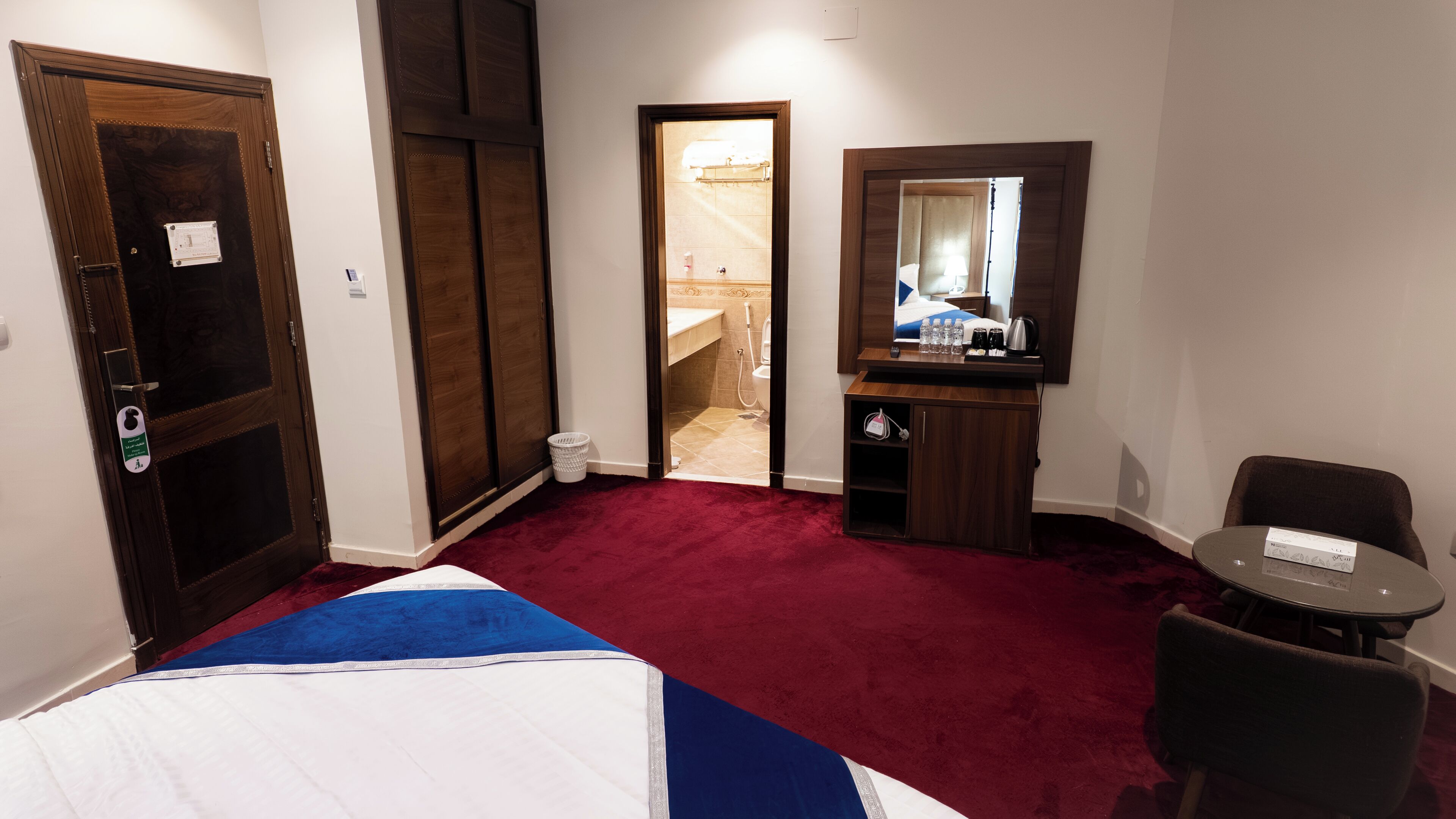 Habitación de hotel con cama, escritorio pequeño, silla, espejo y baño visible a través de una puerta abierta.